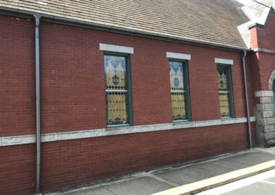Red brick church side. Three windows framed in green. Two downspouts in this image connecting to teh underground pipes in the sidewalk.
